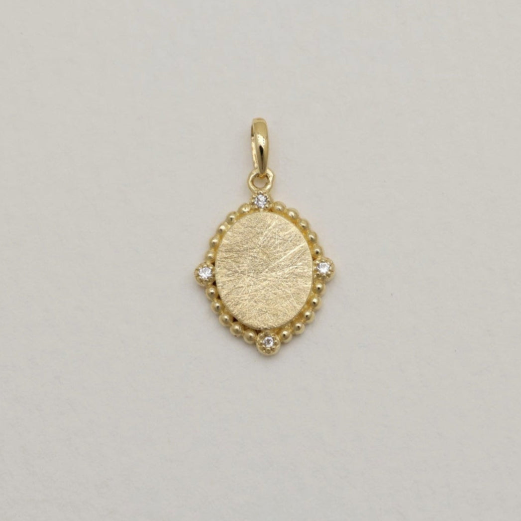 14K gold charm pendant with oval brushed center and four cubic zirconia, surrounded by beaded details.