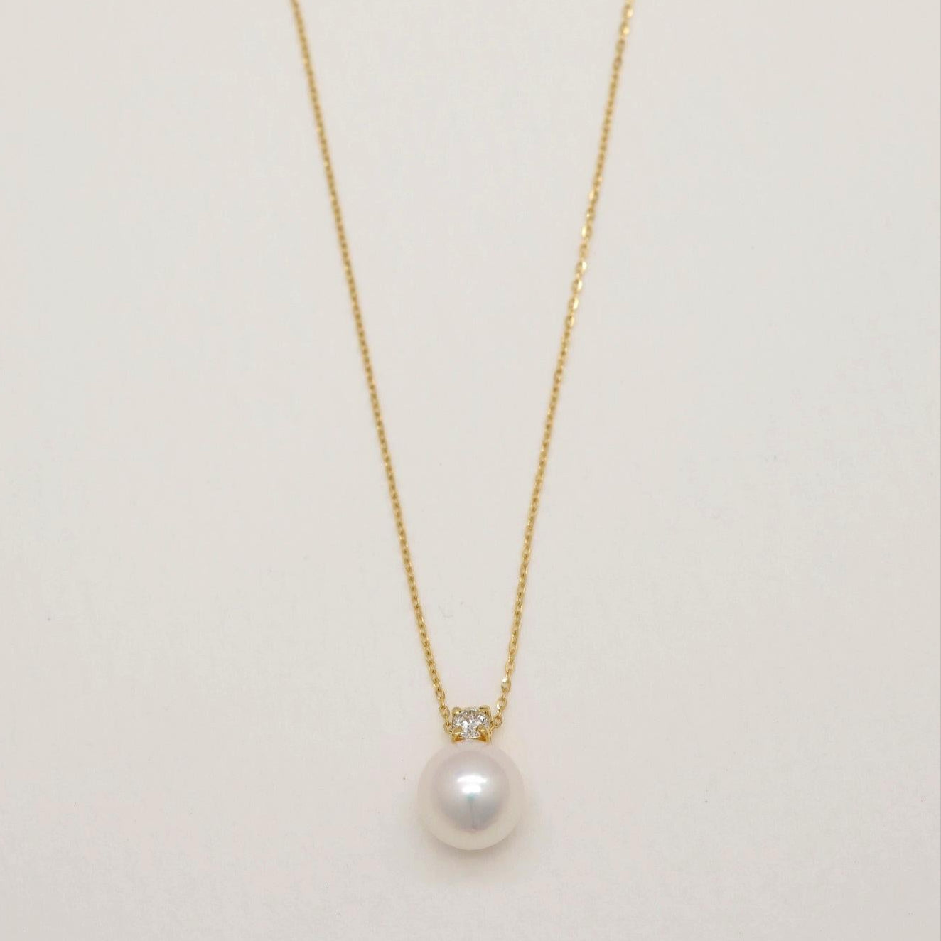 Close-up of 18K gold necklace with Akoya pearl and natural diamond pendant on a light background.
