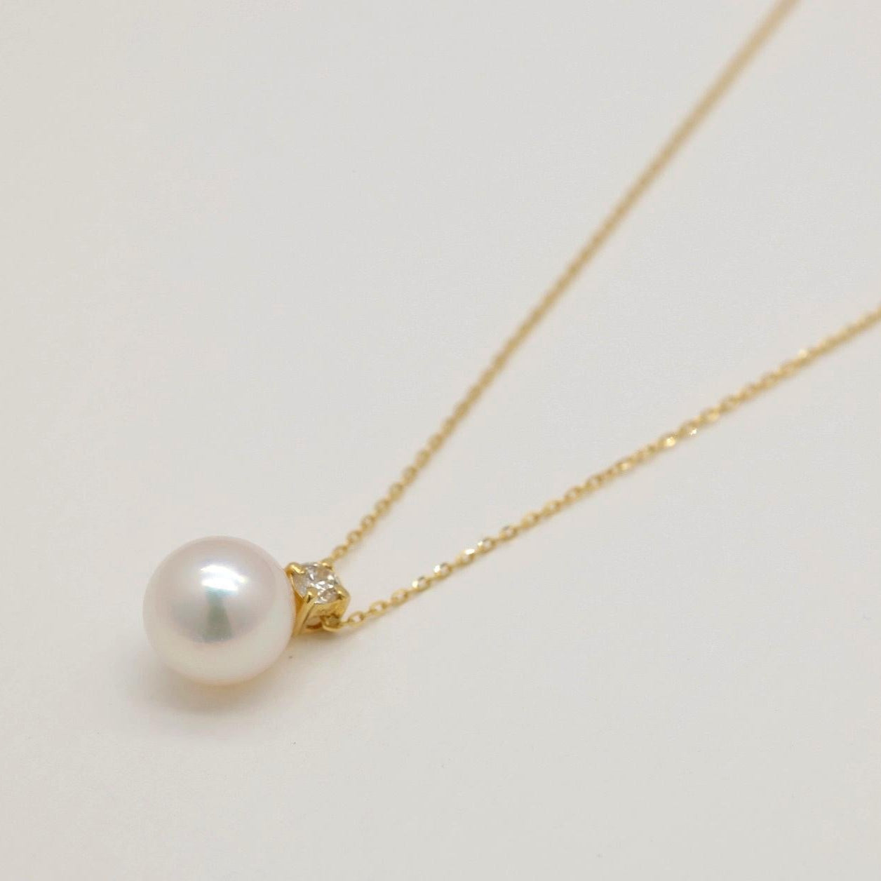 Close-up of 18K gold necklace with Akoya pearl and natural diamond pendant on a light background.