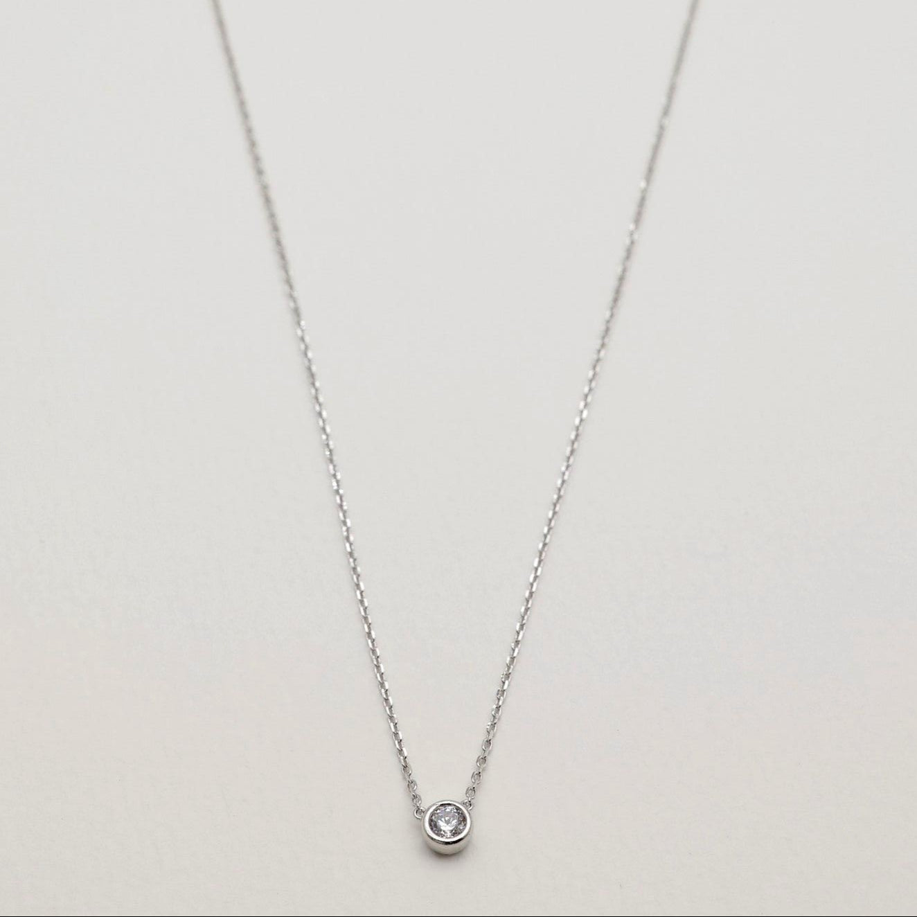 Full view of a dainty silver necklace with a bezel-set cubic zirconia stone, arranged in a delicate curve on a pale surface.