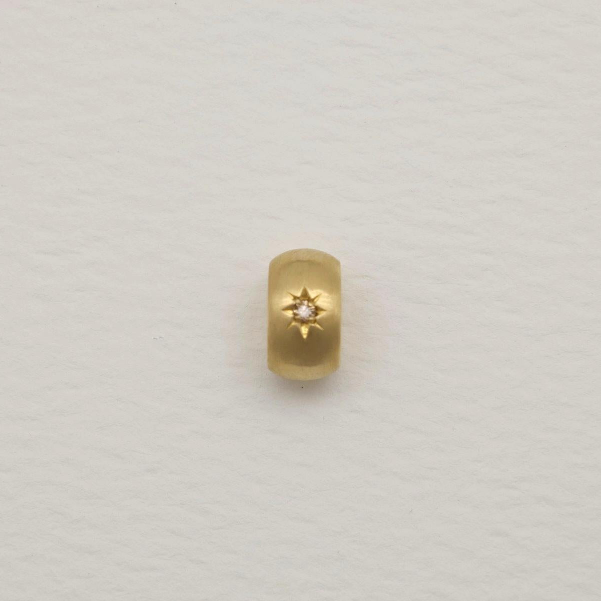 Gold oval pendant with a matte finish and engraved starburst set with a tiny diamond, photographed on a soft white background.