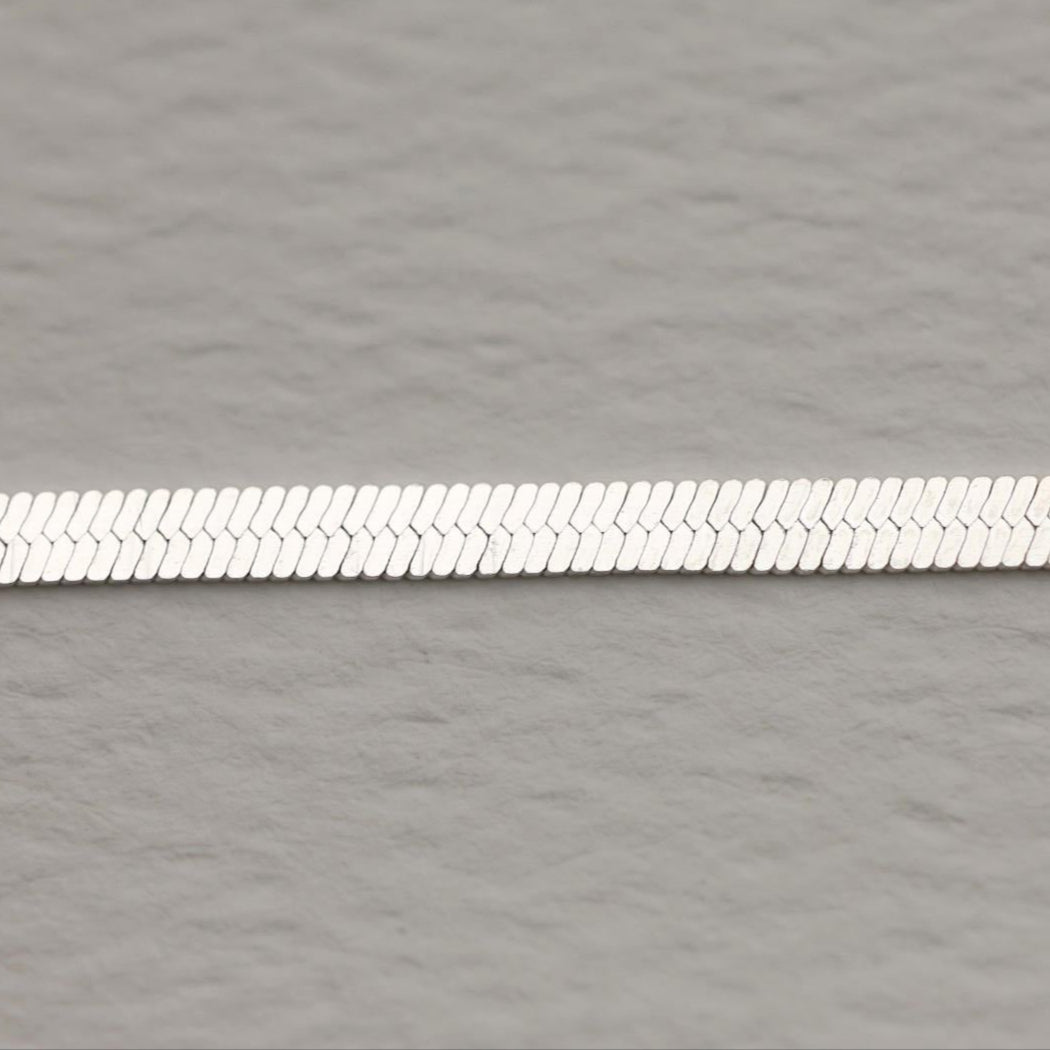 Flat herringbone bracelet in polished 925 sterling silver with extender chain, shown on a soft grey background.