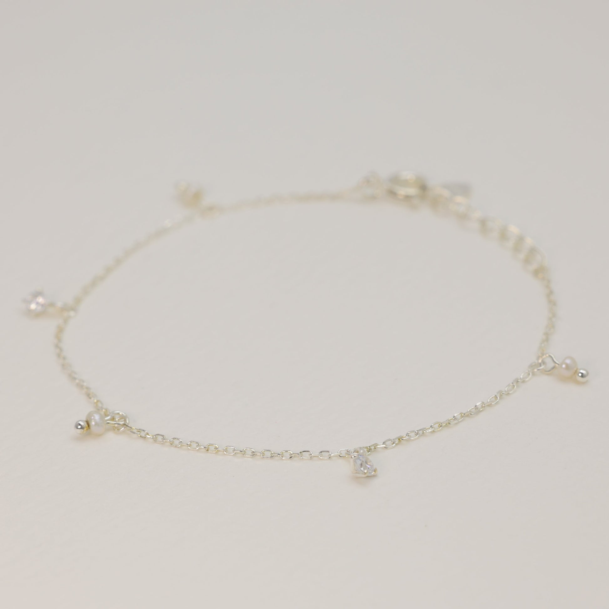 Side view of the sterling silver charm bracelet showing fine chain detail and small dangling pearl and zirconia charms, softly lit on a cream surface.

