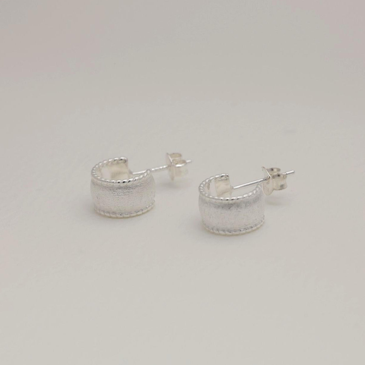 Sterling silver huggie earrings with a wide textured band and polished beaded edge, softly sculpted with matte and glossy contrast.