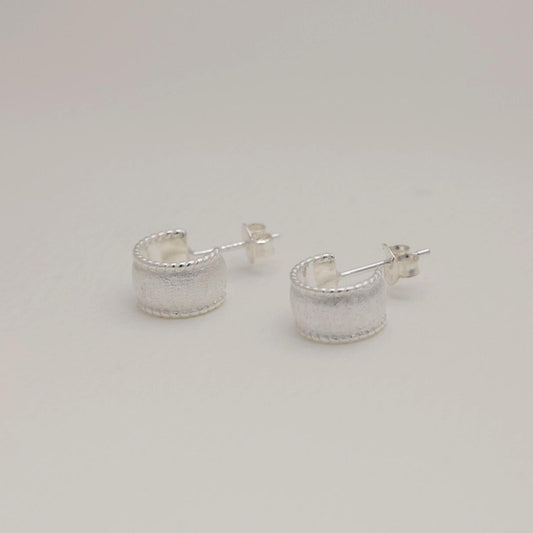 Sterling silver huggie earrings with a wide textured band and polished beaded edge, softly sculpted with matte and glossy contrast.