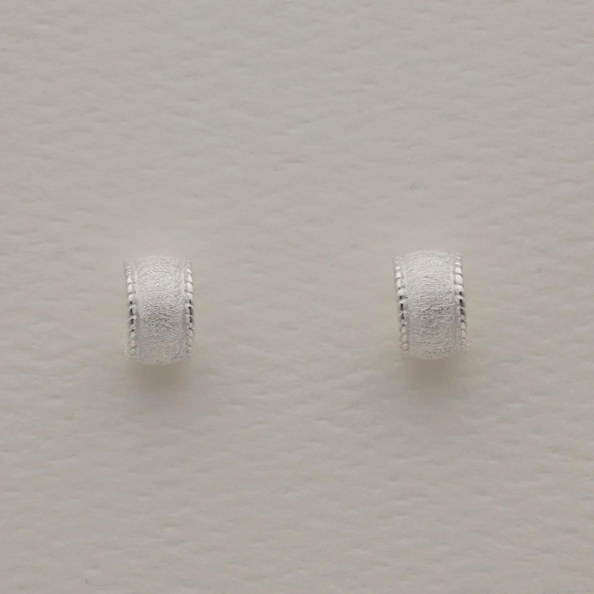 Sterling silver huggie earrings with a wide textured band and polished beaded edge, softly sculpted with matte and glossy contrast.