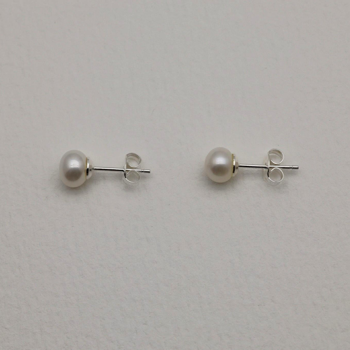 Close-up of 925 sterling silver pearl stud earrings on textured neutral background, showing round natural pearls with minimalist push-back posts.