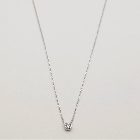 Full view of a dainty silver necklace with a bezel-set cubic zirconia stone, arranged in a delicate curve on a pale surface.