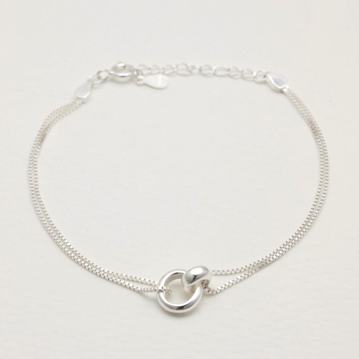 Sterling silver dual-chain bracelet with sculptural ring detail, laid flat on a cream background.
