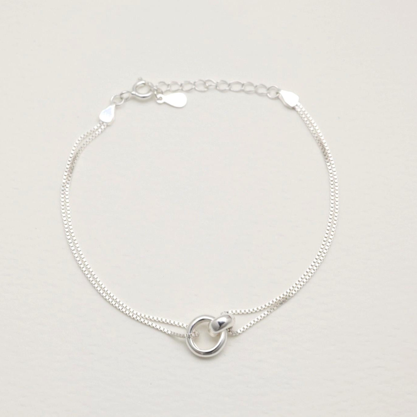 Sterling silver dual-chain bracelet with sculptural ring detail, laid flat on a cream background.