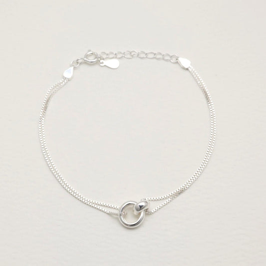Sterling silver dual-chain bracelet with sculptural ring detail, laid flat on a cream background.