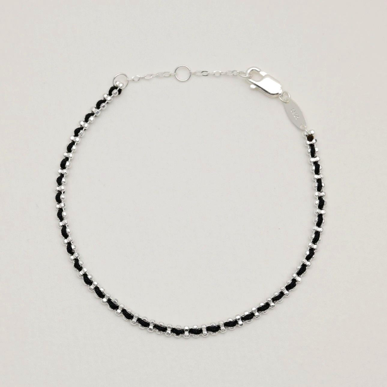Flat lay of the Agnès Woven Bracelet in sterling silver with black thread detail on a neutral background.