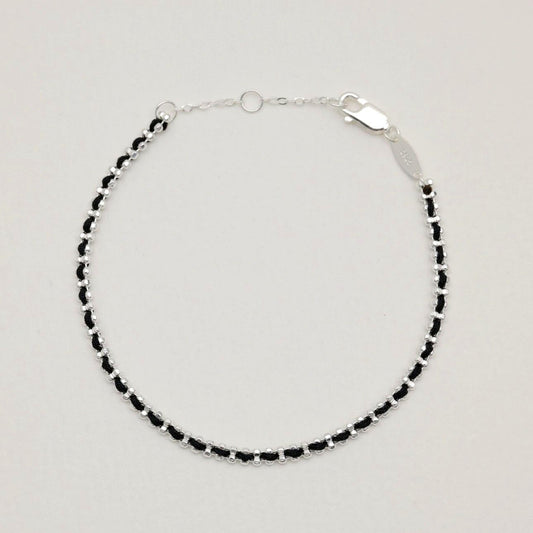 Flat lay of the Agnès Woven Bracelet in sterling silver with black thread detail on a neutral background.